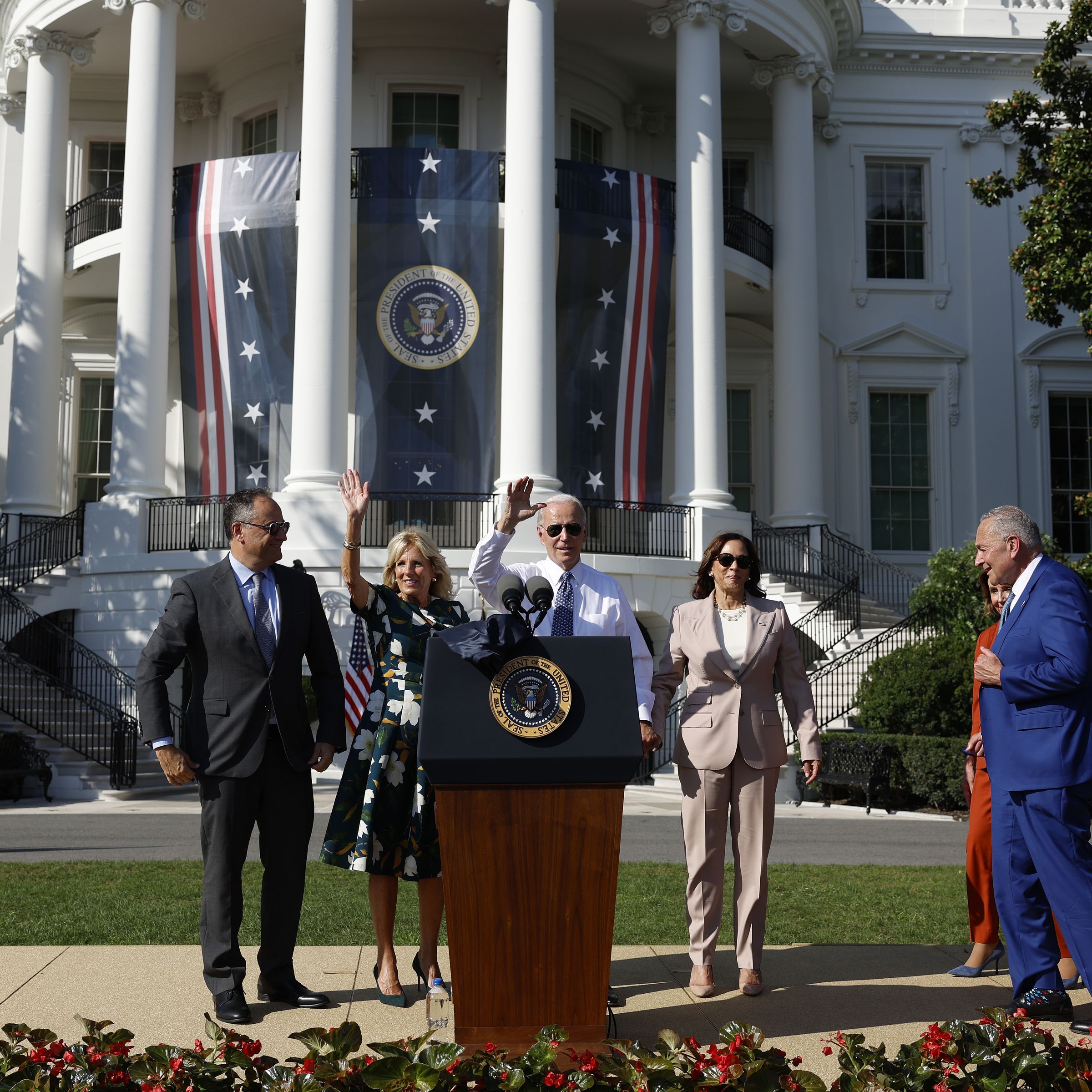 Joe Biden stands at a podium in front of the White House with people standing behind him.