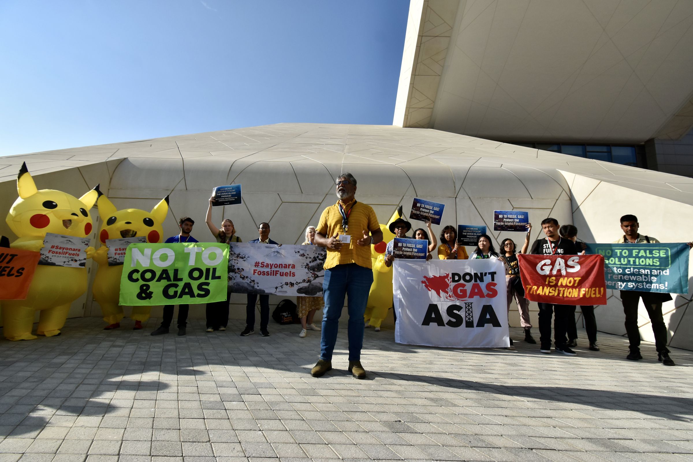 Three people wearing inflatable Pikachu stand among a crowd protesting fossil fuels. One person in a costume holds a sign that says “#Sayonara FossilFuels”. Other demonstrators hold banners that say “No to coal, oil &amp; gas” and “Don’t Gas Asia”.