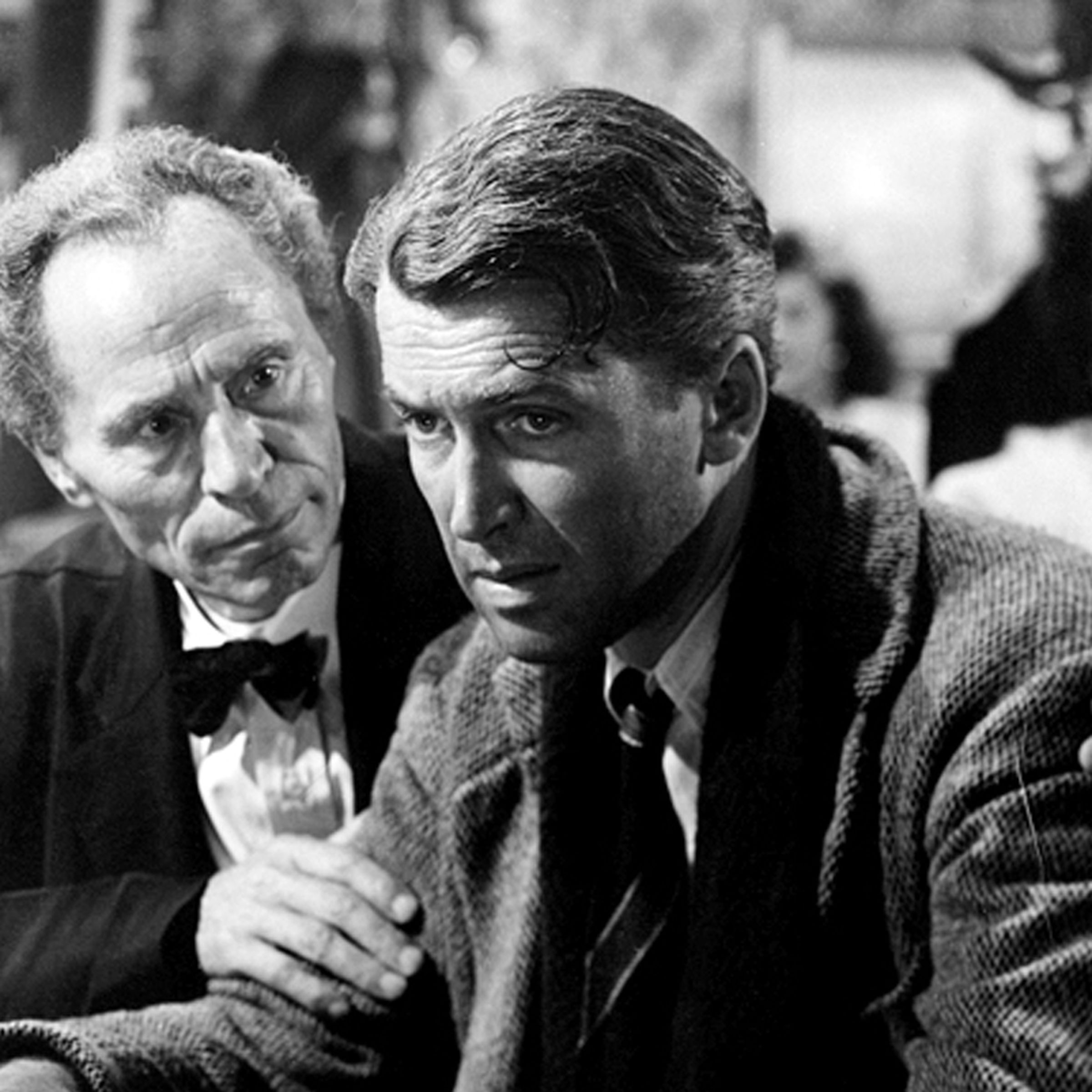 A man in a wool jacket and a scarf being comforted by another man in a wool jacket and a bowtie. Both men are standing at a brightly-lit bar or at a restaurant.
